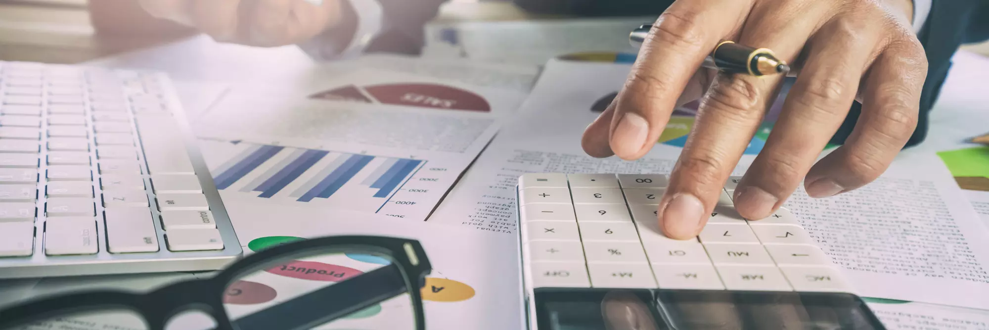 Amministratore di sostegno. Primo piano di una mano che utilizza una calcolatrice su documenti aziendali, inclusi grafici e tabelle. Dietro, occhiali e fogli di analisi finanziaria.