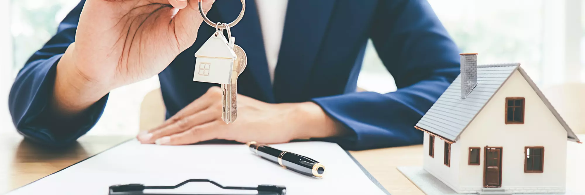 Contratto affitto. Una persona in giacca che tiene chiavi a forma di casa e un'altra chiave, appoggiata su un tavolo, vicino a una penna e un modello di casa.