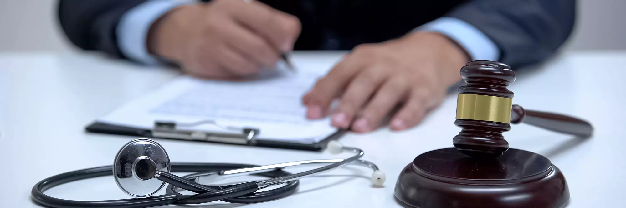 Responsabilità medica. Un avvocato scrive su un documento legale, con un martello di giustizia e uno stetoscopio sul tavolo, simboleggiando la connessione tra legge e salute.