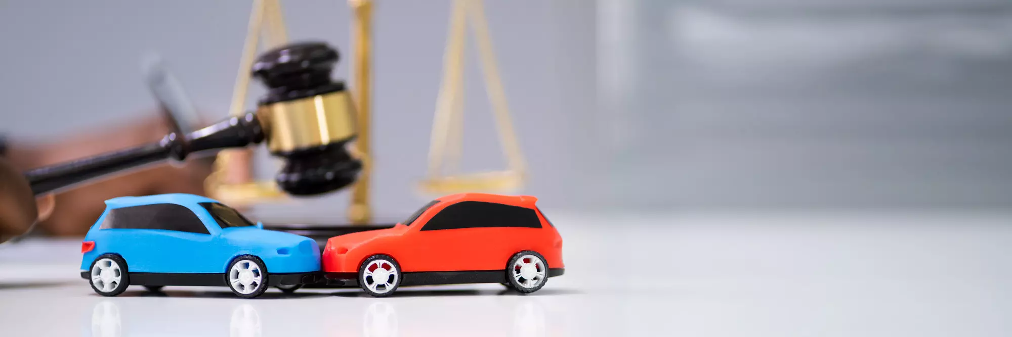 Risarcimento danni stradali. Due piccole auto giocattolo, una rossa e una blu, si trovano in primo piano, mentre sullo sfondo si intravedono una bilancia di giustizia e un martello del tribunale, simbolizzando il tema legale.
