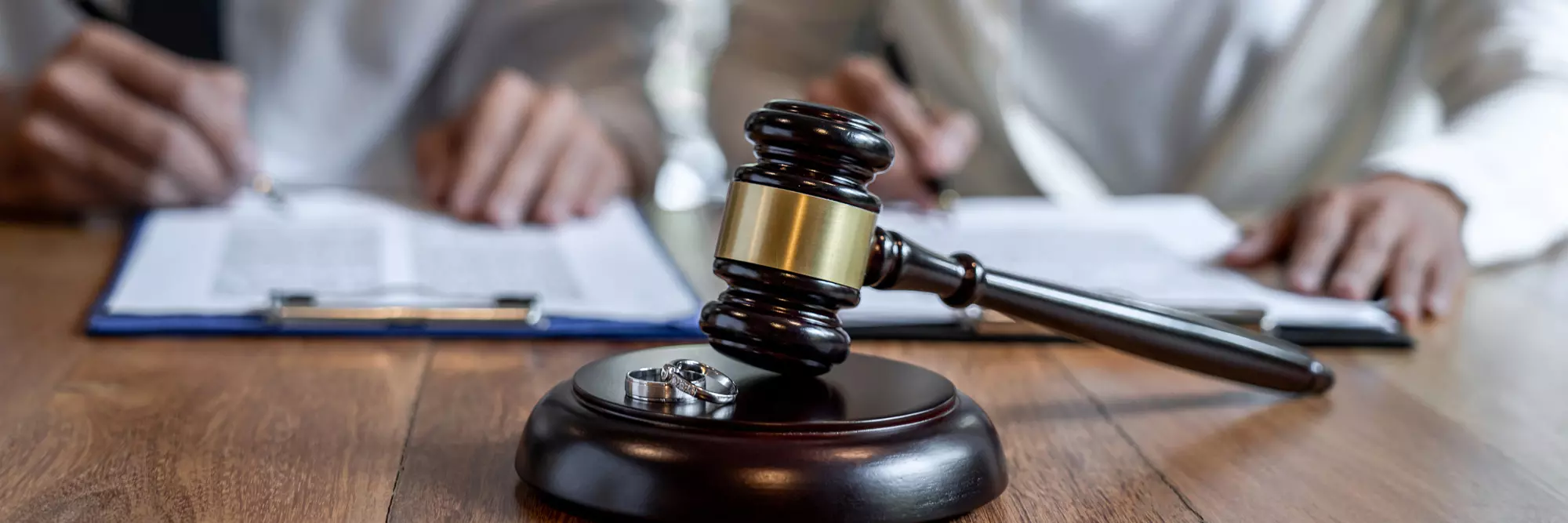 Separazione e divorzi. Martello del giudice su un tavolo di legno, con due fedi nuziali affianco e una persona che scrive documenti. Atmosfera legale.
